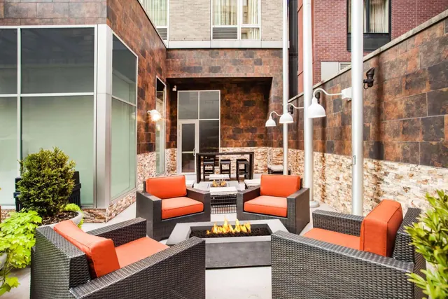 An outdoor courtyard with wicker lounge chairs with orange cushions around a gas fire pit, with brick and stone walls and white wall lamps.