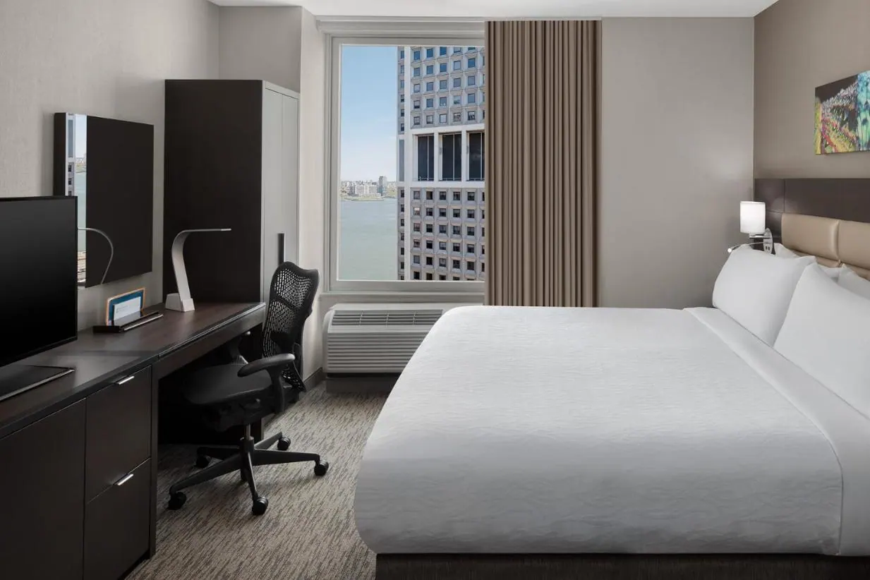 A bedroom with a king bed, a desk with a chair and television, and a window looking over water toward a tall building.