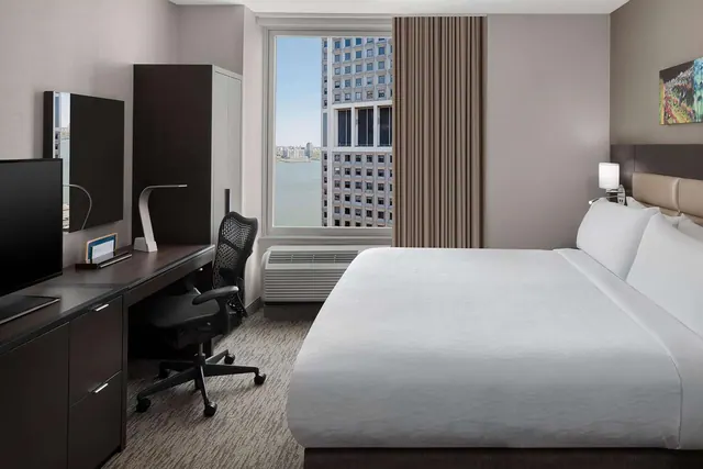 A hotel bedroom with a king bed, a work desk with an office chair, a wall-mounted television, and a window overlooking a river and city buildings.