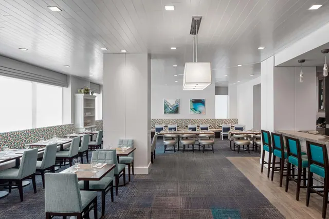 A restaurant dining room with teal upholstered chairs at wooden tables, a counter with bar stools on the right, and a large pendant light overhead.