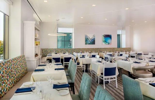 A bright dining room with white-clothed tables, navy napkins, teal chairs and a multicolored dotted banquette along the wall.