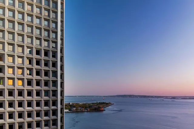 A view over the harbor at dusk, with a high-rise facade on the left, open water, a small fortified island and a distant shoreline.