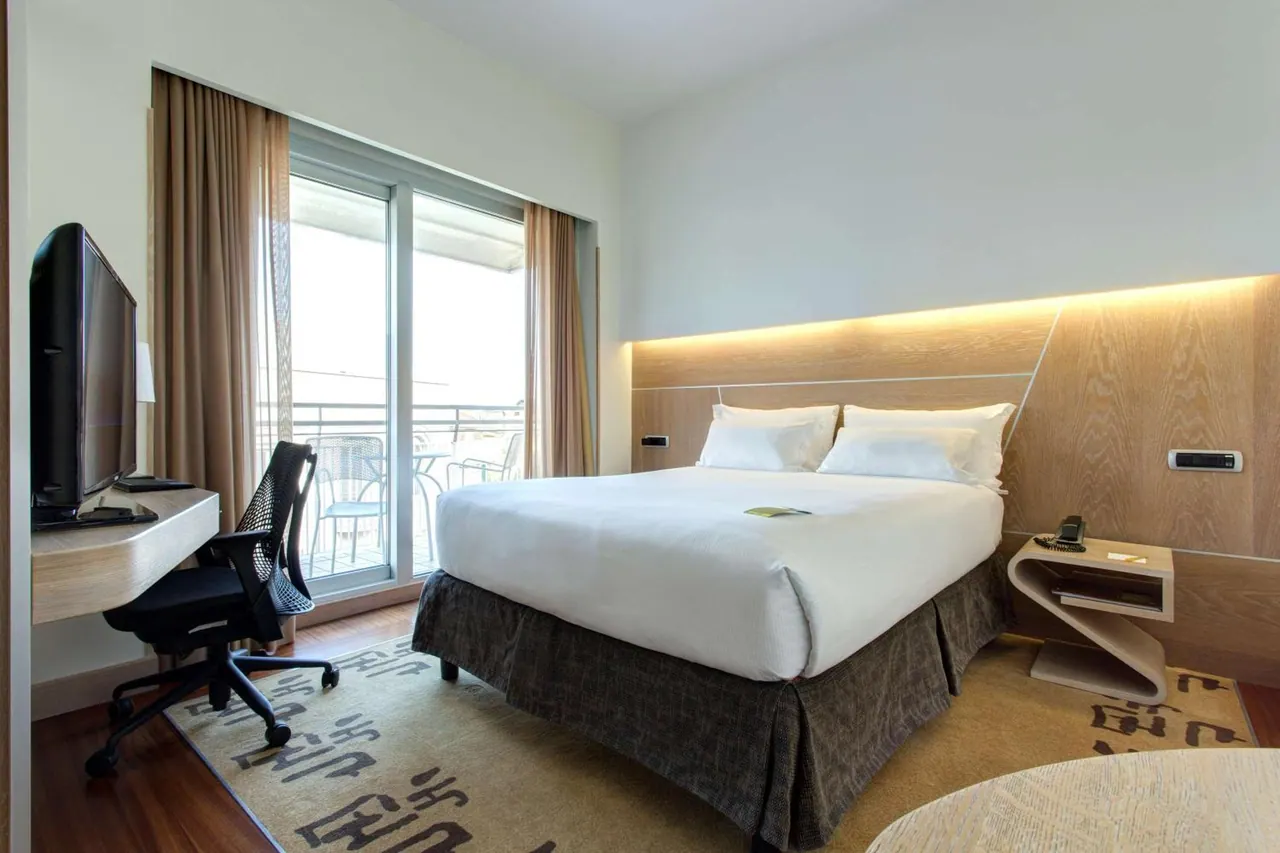 A bedroom with a made double bed, a wood-panelled headboard with warm lighting, a desk with a television and chair, and a glass door opening onto a balcony with metal chairs.