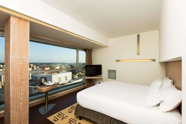 A bedroom with a made double bed, a wall-mounted television, a kettle on a round side table, and floor-to-ceiling windows looking over rooftops and buildings.