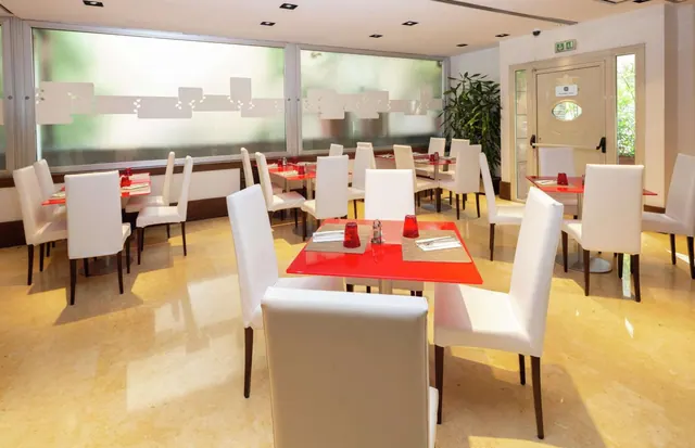 A dining room with red-topped tables, white upholstered chairs, frosted windows, a potted plant and a door with an illuminated exit sign.
