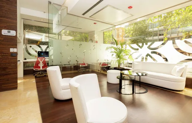 A hotel lounge with white armchairs, a white sofa and glass coffee tables sits beside a glass-walled business center, with a large window looking onto trees and buildings outside.