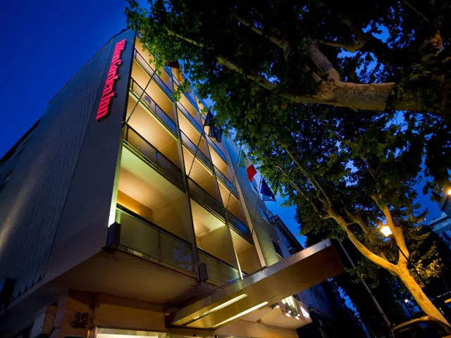 Night exterior of a multi-storey hotel with an illuminated red sign on its facade, a lit canopy at street level, flags on poles and a large tree in the foreground.