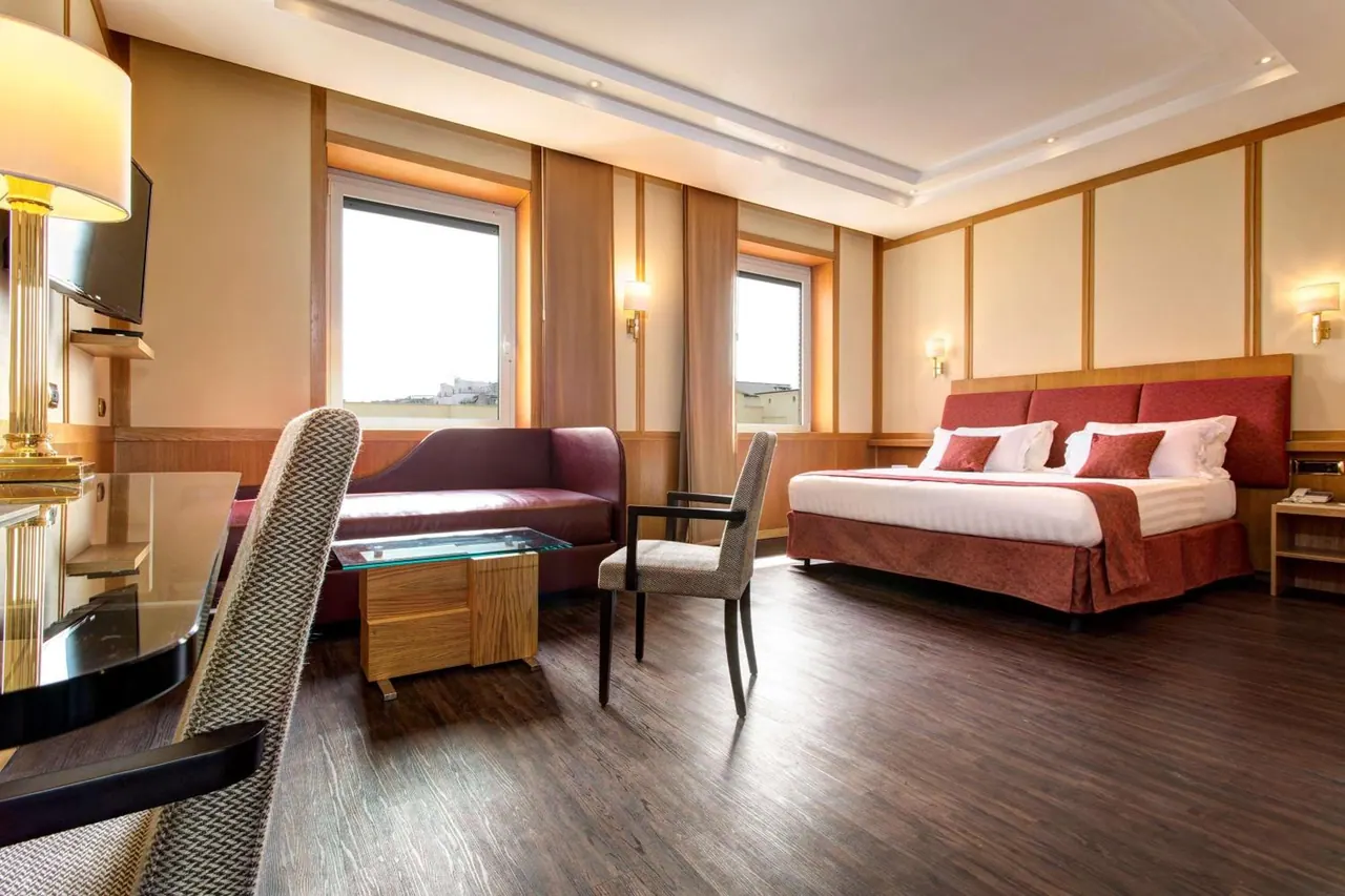 A hotel bedroom with a king bed with a red headboard, a sofa, a chair and desk, and a wall-mounted television, with two windows showing buildings outside.