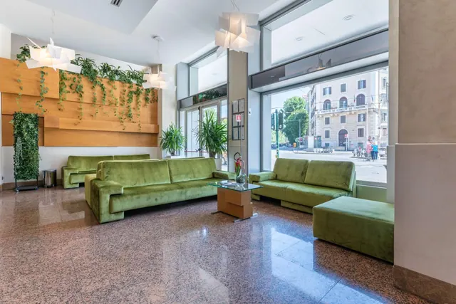 A lobby seating area with green velvet sofas, a glass coffee table and a polished terrazzo floor, with large windows looking onto a street with a building and pedestrians outside.