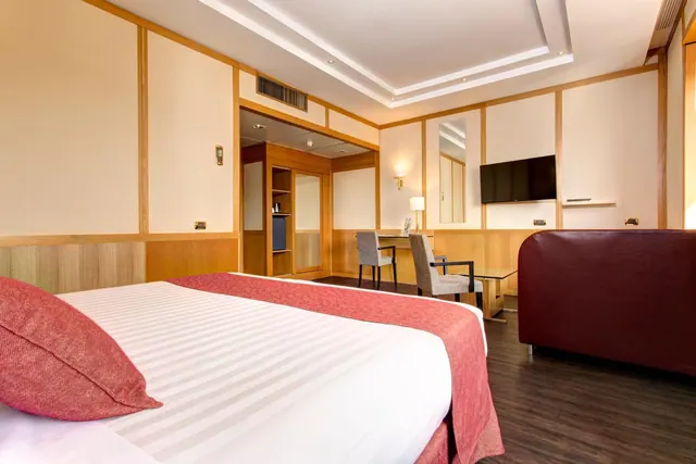 A double bed with a red throw and pillow in a wood-panelled room, with a wall-mounted television, a desk with two chairs and a leather sofa.