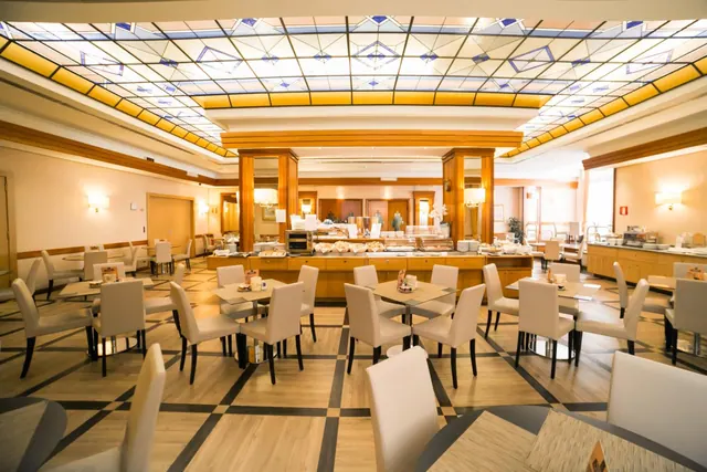 A breakfast dining room with a stained glass ceiling, rows of white chairs and tables, and a buffet counter at the back.