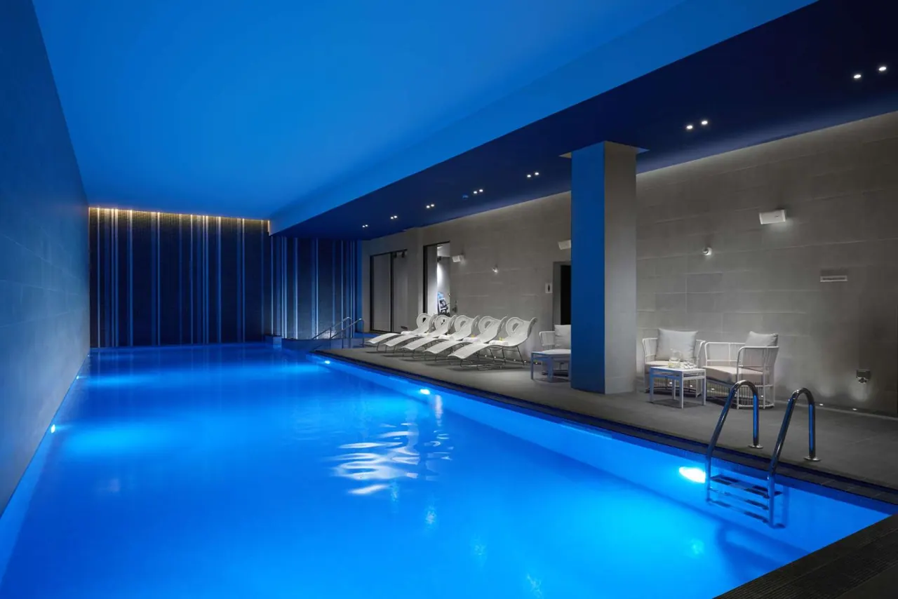 An indoor pool lit in blue, with white sun loungers and seating along the side under ceiling spotlights.