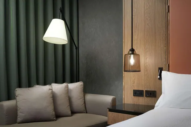 A hotel bedroom corner with a grey sofa and cushions, a floor lamp beside green curtains, a pendant lamp over a bedside table, and the edge of a made bed.