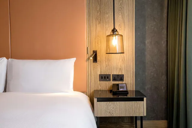 A made bed with white linen sits beside a wood-panelled bedside table holding a telephone, under a glass pendant lamp.