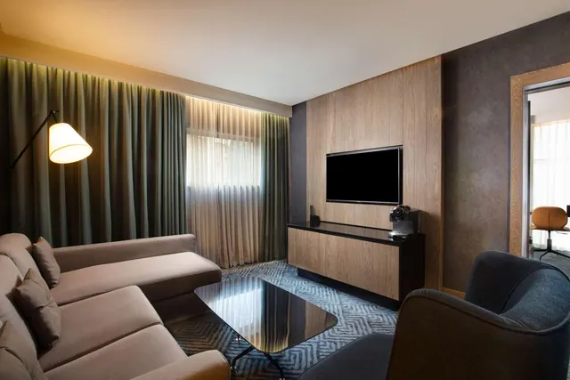 A hotel living area with a grey sofa, a wall-mounted television above a wood sideboard with a coffee machine, a floor lamp, and sheer curtained windows.