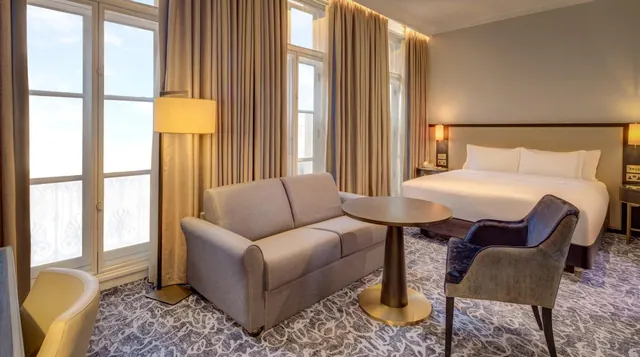 A hotel bedroom with a king bed, a grey sofa, a round wooden table and an armchair beside tall windows with beige curtains.