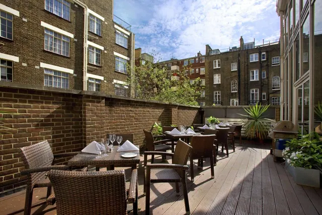 An outdoor wooden-decked terrace with wicker chairs and laid tables, enclosed by a brick wall and overlooking surrounding brick buildings and trees.