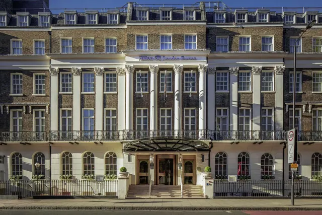 The front facade of a multi-storey brick hotel building with white pilasters, a canopied entrance with steps, and a branded sign above the doors, viewed from the street.