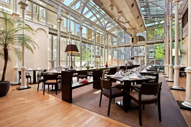 A glass-roofed restaurant with white columns, laid dining tables and a potted palm, with brick buildings visible through the windows.
