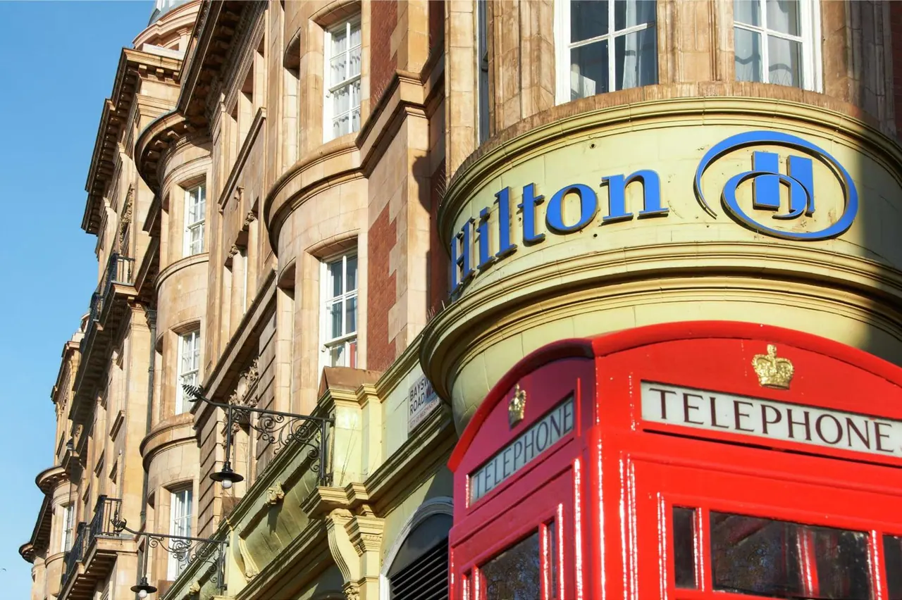 The rounded corner of a Hilton hotel building with its blue logo sign, beside a red British telephone box under a clear blue sky.
