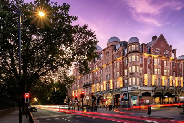 Dusk view of a red brick hotel building with illuminated windows and a Hilton sign, with traffic light trails on the street below.