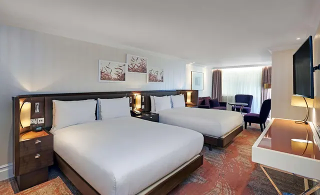 A hotel bedroom with a pair of double beds, a wall-mounted television, a desk, and purple armchairs by a window.