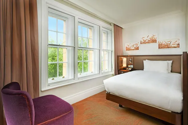 A bedroom with a double bed with white linen, a purple armchair, and tall sash windows looking out onto trees.