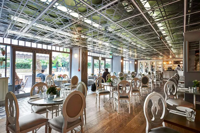 A dining room with rows of tables and oval-backed chairs under a patterned metal ceiling, with windows onto a street.