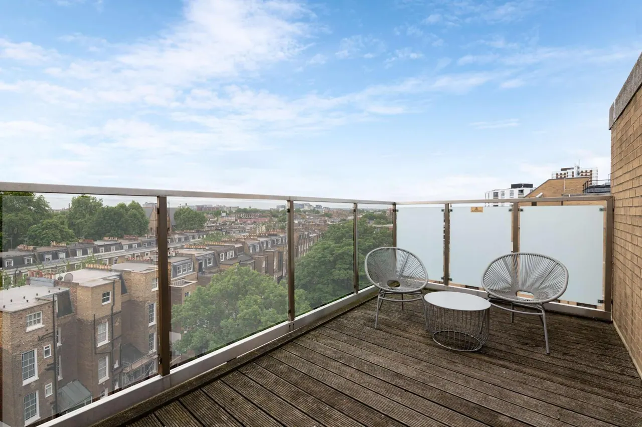 A private balcony with glass railings, two wicker chairs and a small round table overlooks terraced houses and trees under a partly cloudy sky.