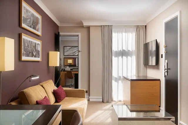 A living area with a tan sofa and red cushions, framed prints on a plum wall, a wall-mounted television, a wooden cabinet, and a desk visible through an open doorway.