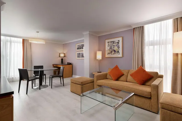A hotel suite living area with a tan sofa, orange cushions, a glass coffee table, a round dining table with four chairs, and tall windows with sheer curtains.