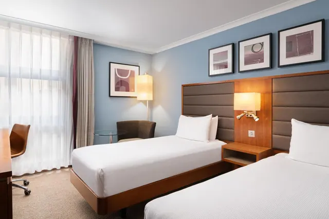 A twin bedroom with two single beds, a wooden desk and chair by the window, and an armchair beside a round glass table.