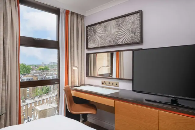 A hotel bedroom with a glass-topped desk, a wall-mounted television and a window overlooking terraced rooftops and trees under a partly cloudy sky.