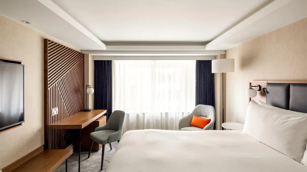 A hotel bedroom with a large bed, a wooden wall-mounted desk with a grey chair, a grey armchair with an orange cushion by the window, and a wall-mounted television.