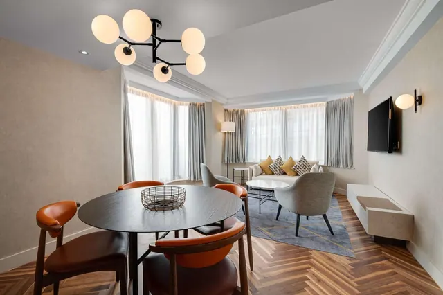 A living and dining area with a round dark table and four orange chairs, a sofa with yellow cushions, an armchair, a wall-mounted television and bay windows with sheer and grey curtains.