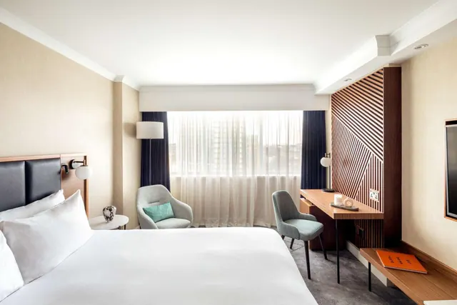 A hotel bedroom with a made white bed, two upholstered armchairs by a window with sheer curtains, and a wooden desk with a chair on the right.