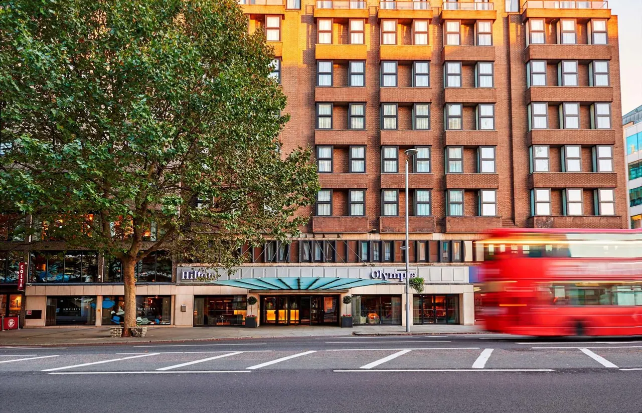 Exterior of a multi-storey brick hotel building with Hilton and Olympia signage above a canopied entrance, a large tree on the left and a blurred red double-decker bus passing on the street.