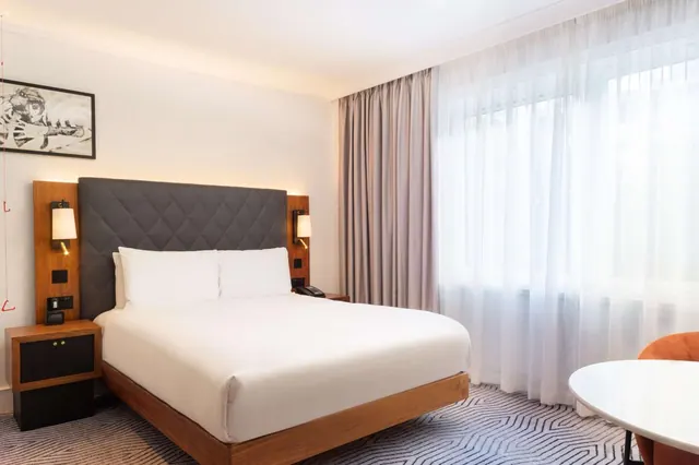 A made-up double bed with a quilted grey headboard and wall-mounted reading lamps sits beside a large window with sheer curtains, and a round white table and orange chair are at the right.