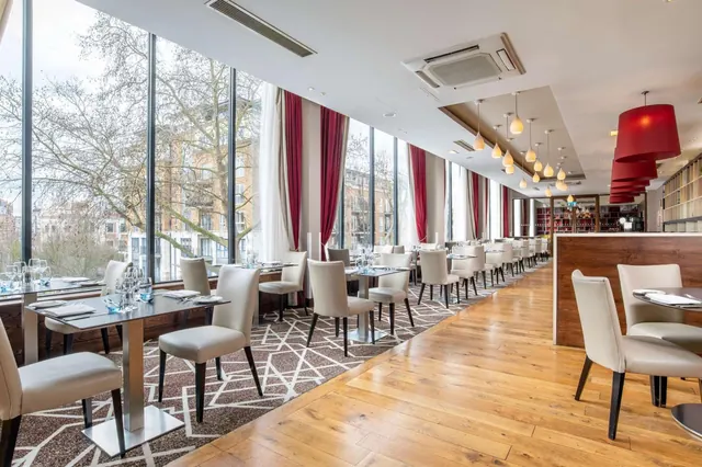 A restaurant dining room with rows of tables and cream upholstered chairs, floor-to-ceiling windows looking out onto bare trees and brick buildings, and red pendant lights.