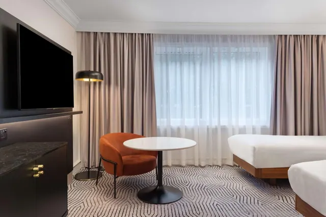 A hotel bedroom with two beds, a wall-mounted television, an orange armchair beside a round white table, and a window with sheer curtains.