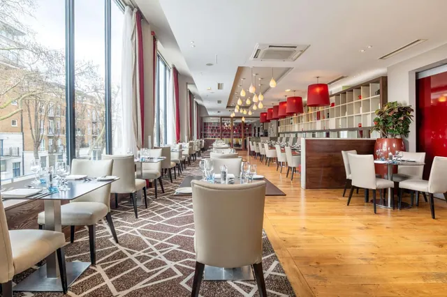 A restaurant dining room with set tables and cream chairs, floor-to-ceiling windows, red pendant lights and a wooden floor.