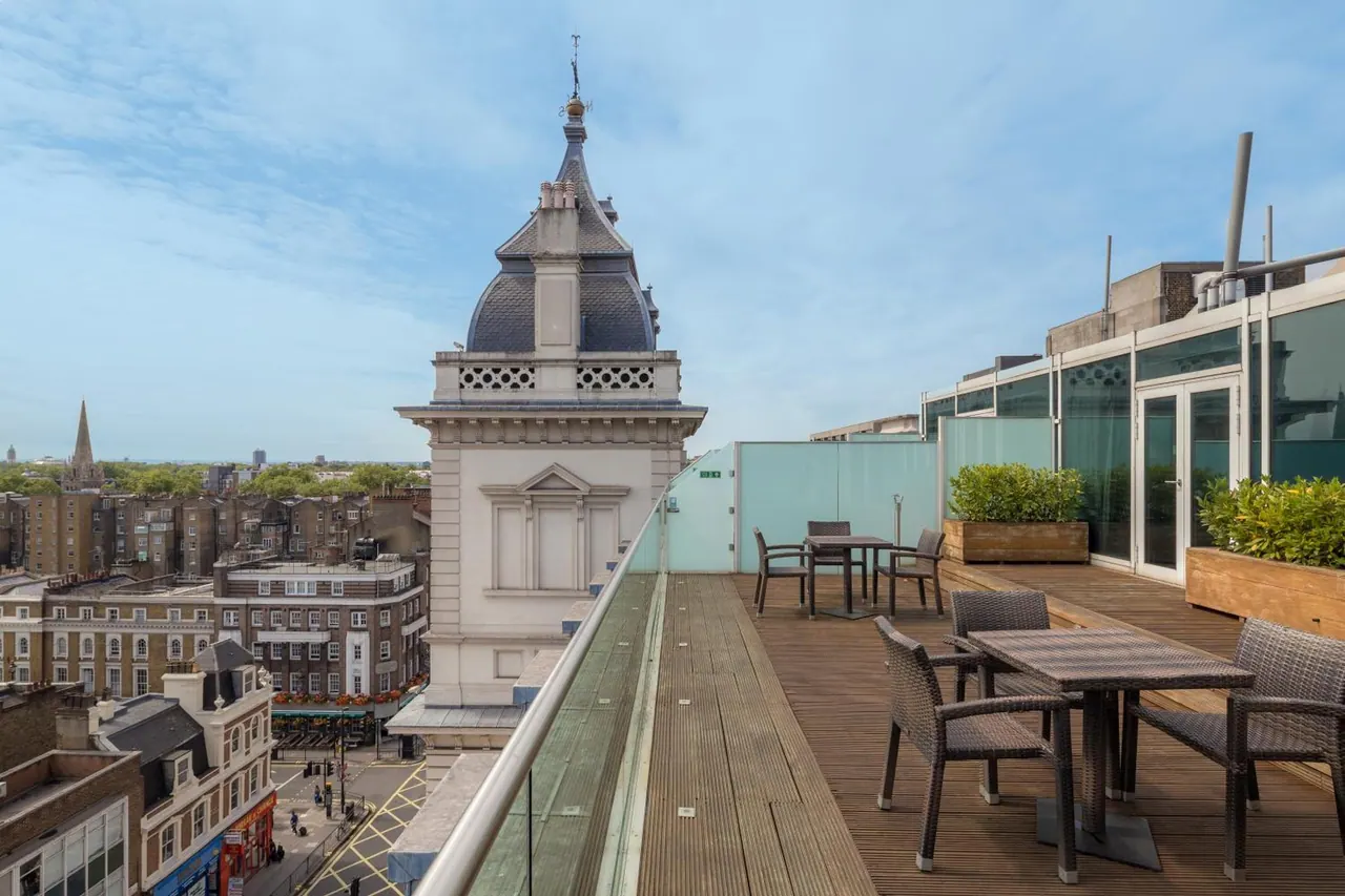 A rooftop terrace with wooden decking, wicker chairs and tables, glass railings and planters, overlooking historic buildings and rooftops under a partly cloudy sky.