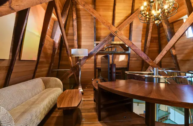 A wood-panelled room with crossed timber beams in the ceiling, a patterned sofa, a floor lamp, a chandelier and a round wooden table beside a glass railing.