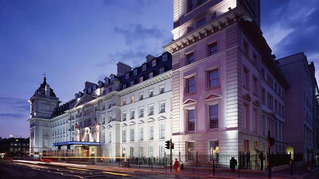 Dusk view of the hotel's lit stone facade and entrance canopy with flags, a street with light trails, and a few pedestrians on the pavement.