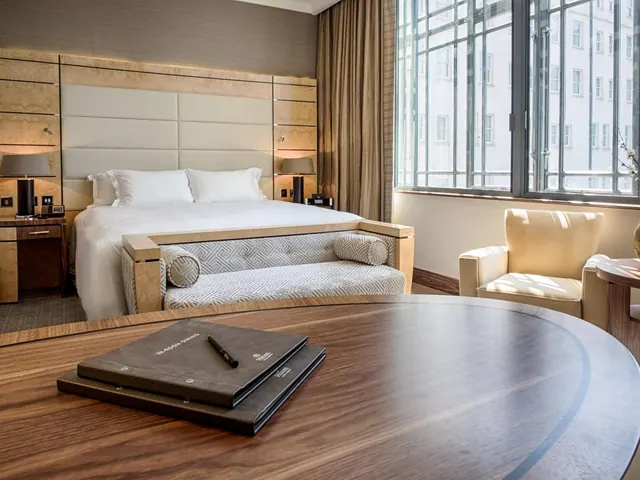 A hotel bedroom with a king bed, an upholstered bench at the foot of the bed, an armchair by the window, and a round wooden table holding two folders and a pen.
