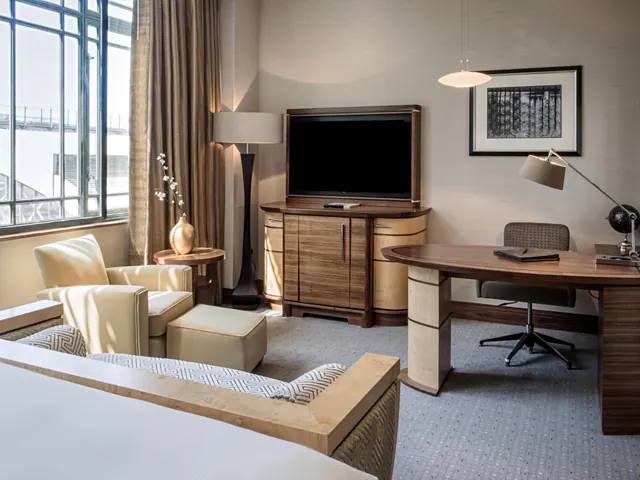 A hotel bedroom with a bed in the foreground, an armchair and ottoman by the window, a television on a wooden cabinet, and a desk with a chair.