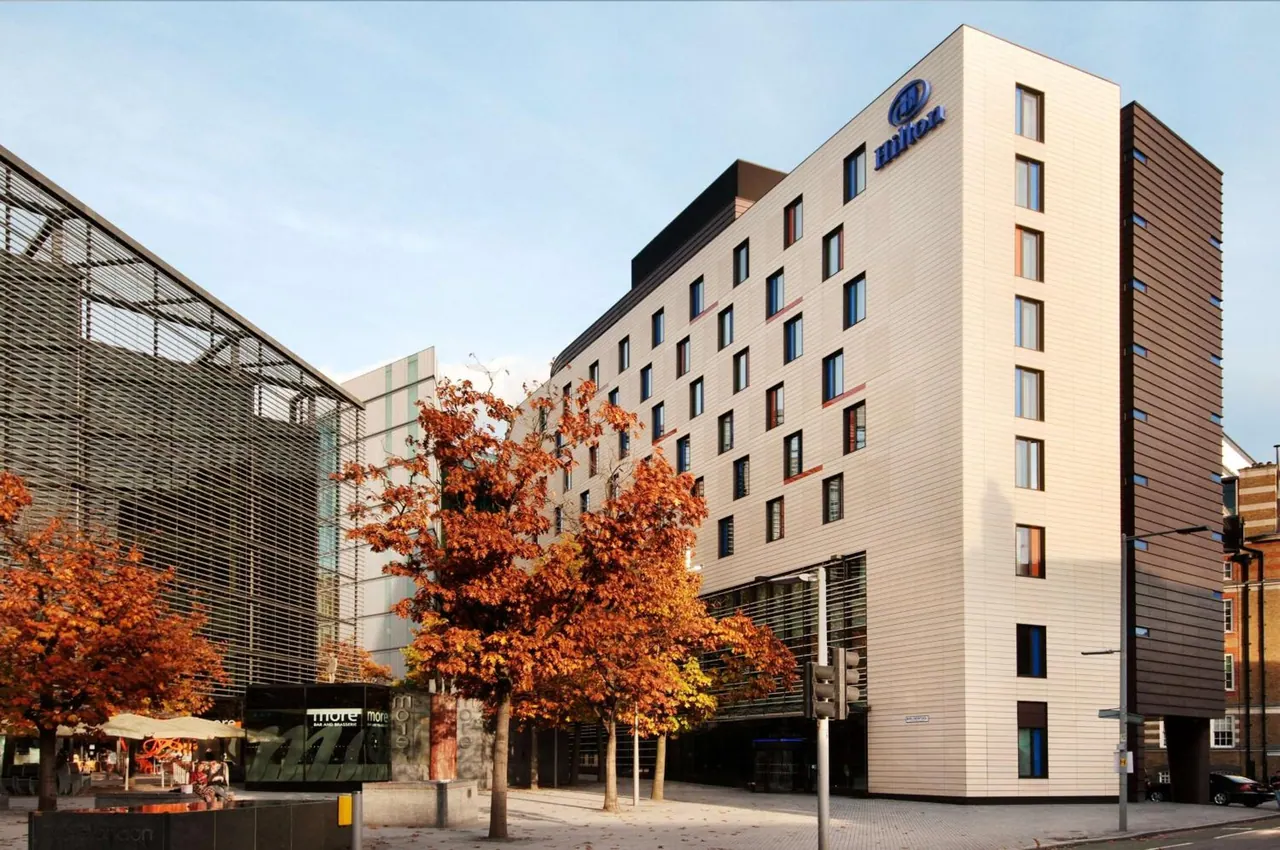 Exterior of a cream-clad Hilton hotel building with autumn trees in front, a traffic light and a parked car on the street.