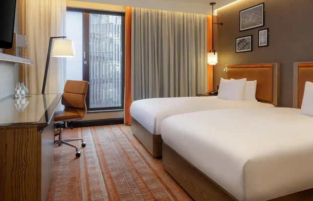 A hotel bedroom with two made beds with white bedding, a wooden desk with a brown chair, a wall-mounted television, and a window looking onto a building.