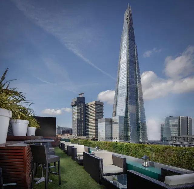 A rooftop terrace with wicker armchairs, high bar tables and potted plants, with The Shard tower rising in the background.