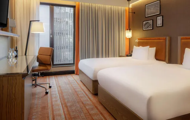 A hotel bedroom with two twin beds, a glass-topped desk with a leather chair, and a window facing a building facade.